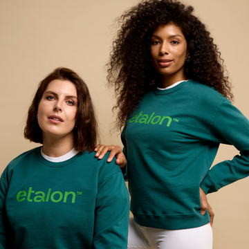 Two women in green eco-friendly Etalon sweatshirts facing forward, each displaying the brand’s front logo design.